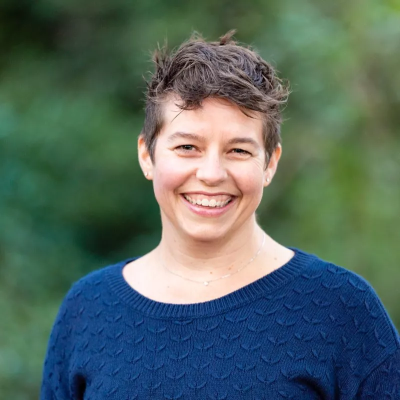 A woman wearing a textured blue sweater is pictured outdoors against a blurred green natural background. The setting appears serene and natural, with soft lighting enhancing the scene. The focus is on the upper body and clothing details.